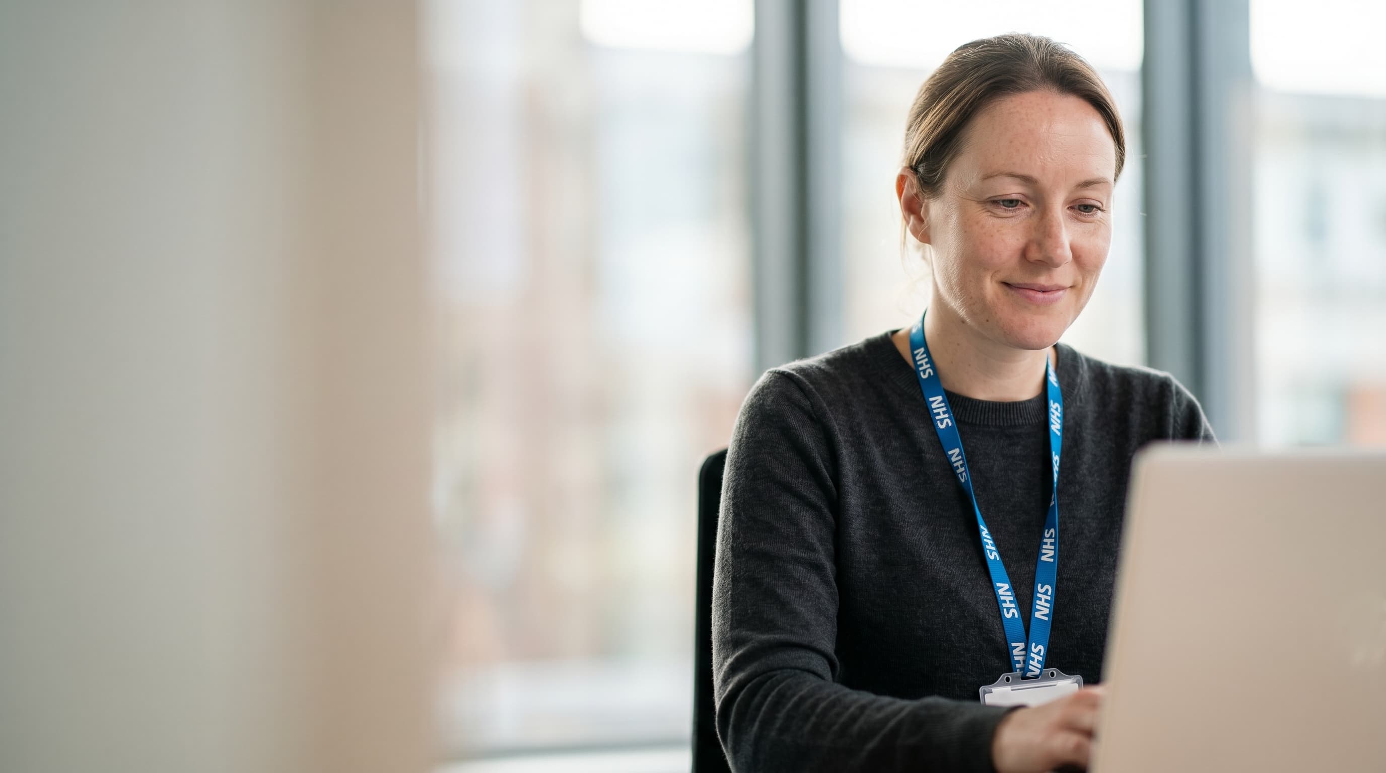 A clinician wearing an NHS lanyard working at a laptop